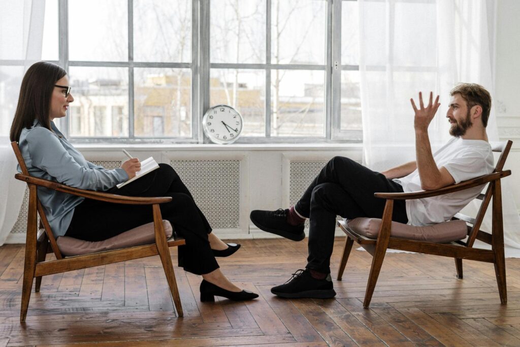 A therapist and patient are seated in chairs in front of a large, bright window discussing rehab