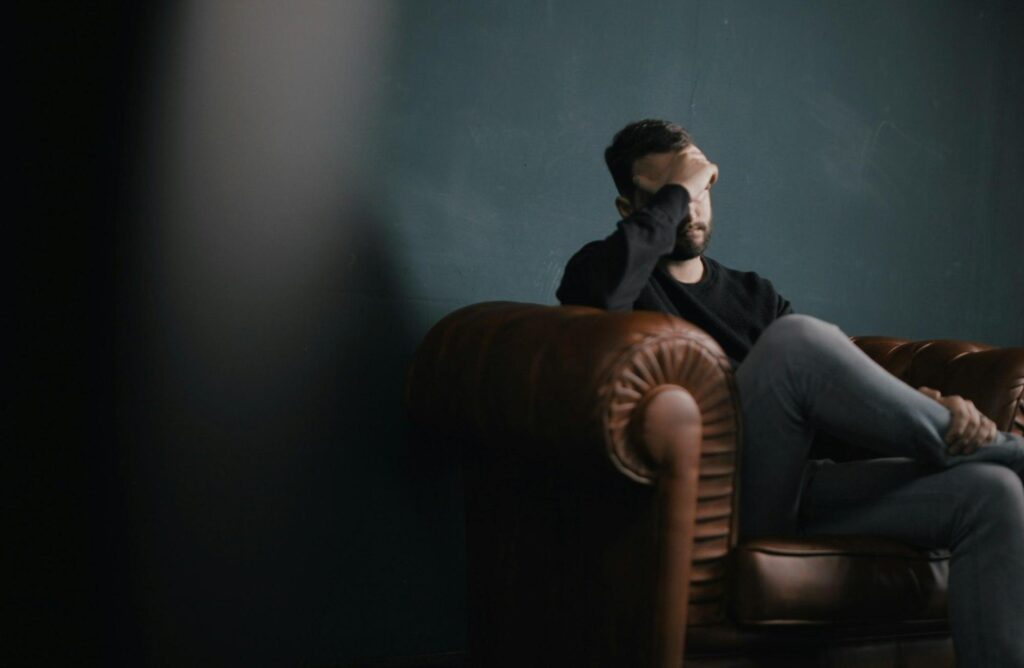 A white man sits with his hand over his head on a brown leather couch, looking depressed.