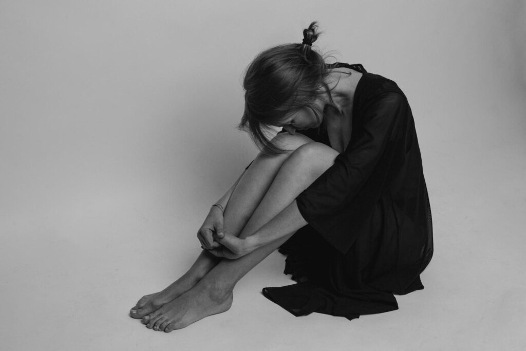 A young woman sits with her arms around her ankles, her head tucked into her knees, and she looks depressed.