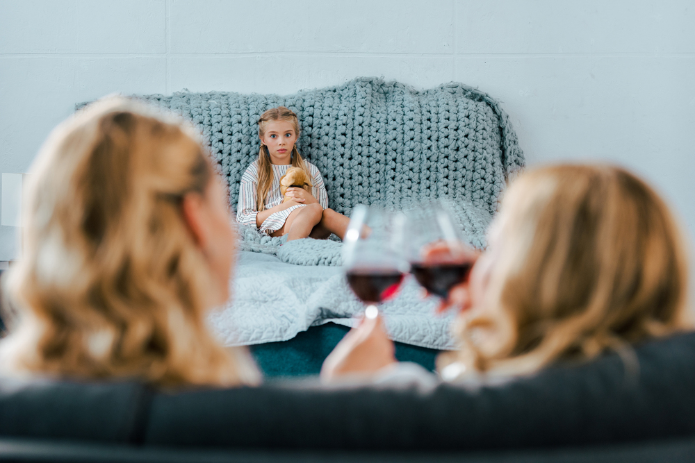 Lonely child sitting on bed with toy while alcoholic mother drinks wine with a friend, showing an example of who would develop an alcohol addiction in adulthood